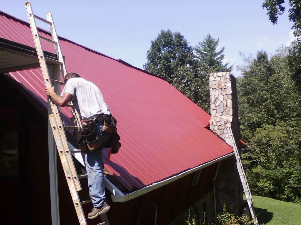 Roofer on a ladder beside a red metal roof and stone chimney — Soffit & Fascia Repair by Roofing Brothers in Bessemer City